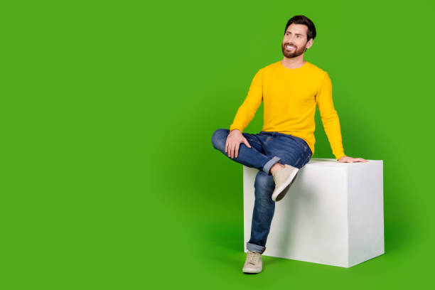 istock Full length photo of handsome young guy look empty space sit relaxed cube dressed stylish yellow garment isolated on green color background 2179529067