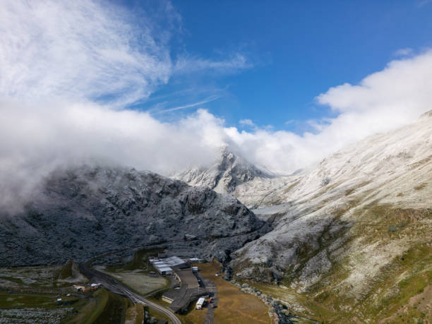 A high-altitude mountain pass with snow-dusted peaks and buildings nestled in the valley below. The clear blue sky and misty clouds create a dramatic and serene landscape.