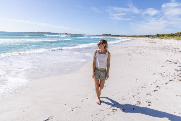A solitary woman strolls along a pristine, white sandy beach with turquoise waters and clear blue skies on a sunny day.