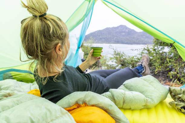 A woman relaxes inside her tent, enjoying a drink while looking out at a serene coastal landscape. The setup includes a cozy sleeping bag and a stunning view of the beach and mountains.