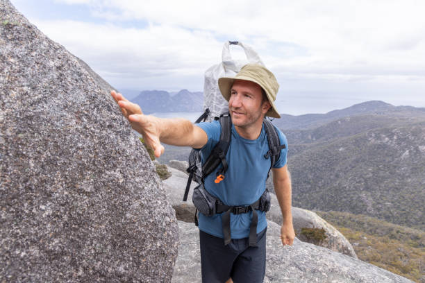 A male hiker, wearing a sun hat and carrying a large backpack, reaches for support while climbing a rocky mountain. The scene features a panoramic view of mountains and coastline under a partly cloudy sky.