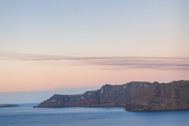 Cette image capture la vue panoramique d'une île de la caldeira située en face du village d'Oia, à Santorini, au coucher du soleil. Les falaises volcaniques imposantes se dressent sur la mer Égée, sous un ciel pastel aux teintes douces et apaisantes. Les formations rocheuses témoignent de l'origine volcanique de l'île, et l'atmosphère sereine reflète la tranquillité d'une soirée d'été sur cette île méditerranéenne emblématique. Le contraste entre les falaises rouges et brunes et le bleu calme de l'eau offre un spectacle naturel impressionnant, parfait pour les amateurs de paysages marins et de nature.