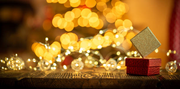 A festive holiday scene featuring a red gift box with a glittery gold lid, surrounded by beautiful bokeh lights. The blurred background shows warm, golden Christmas lights, creating a cozy and enchanting atmosphere.