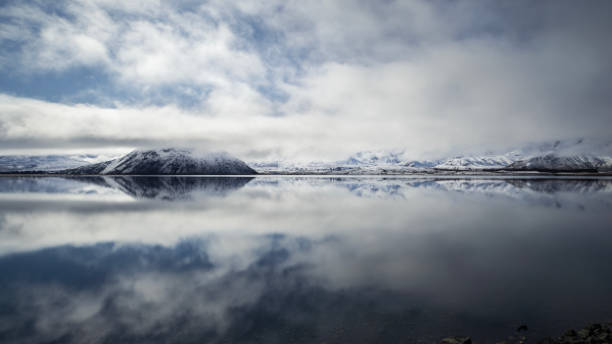 Mid-morning at Lake Heron and the mist is finally starting to clear, revealing a blue sky. The mountains in the distance are bathed in sunlight, making the freshly fallen snow on them shine brightly. The still waters of the lake reflect the scene.\nLake Heron is one of a group of remote lakes called the Ashburton Lakes. They are situated in the mid Canterbury region on New Zealand's South Island.