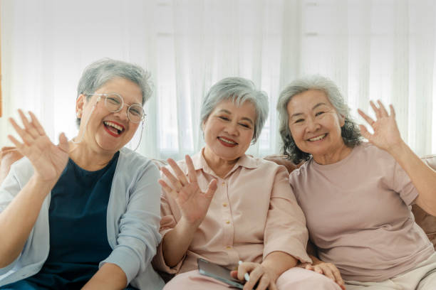 istock Three elderly women sit together, smiling and waving at the camera, radiating joy and friendship. The image emphasizes companionship, happiness, and togetherness in a relaxed, homey setting. 2179658962