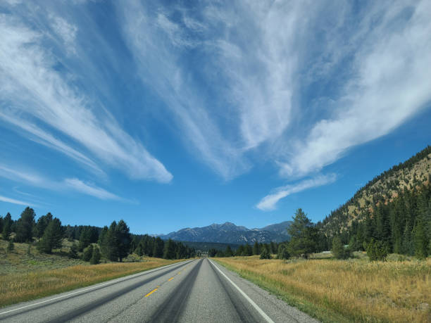 A scenic view of a rural road in the state of Montana, winding through a mountainous landscape under a blue sky with wispy clouds.