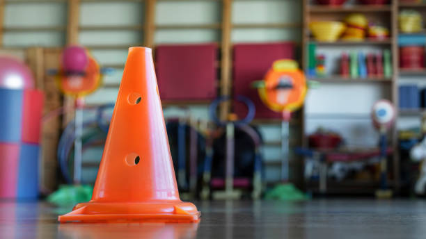 Bright orange plastic cone placed on floor in gym or playroom with colorful equipment blurred in background stock photo