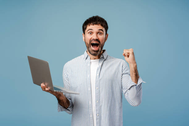 istock Smiling overjoyed bearded man, winner holding laptop, looking at camera, isolated on blue background 2179675937