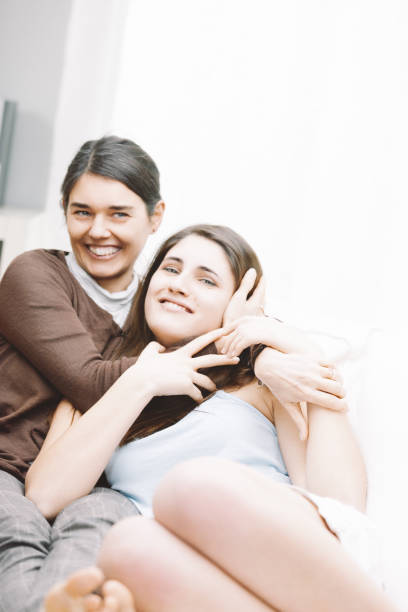 Two young women laughing and hugging on a white couch in a bright living room, radiating joy and togetherness in their cozy home