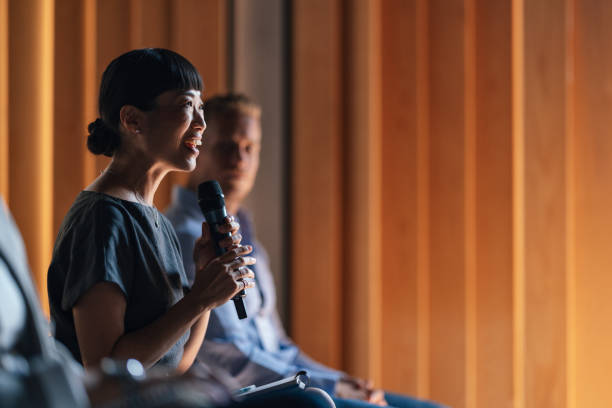 A confident speaker addresses a business conference audience, holding a microphone, with focused engagement. The setting evokes a professional atmosphere suitable for lectures and discussions.