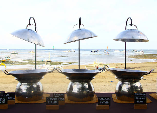 This image features a charming beachfront restaurant, illuminated at sunset. The blue and wooden tables and chairs are set under shade-providing trees and umbrellas, overlooking the calm sea with anchored boats in the background. Ideal for topics related to tropical travel, beach vacations, dining, and relaxation