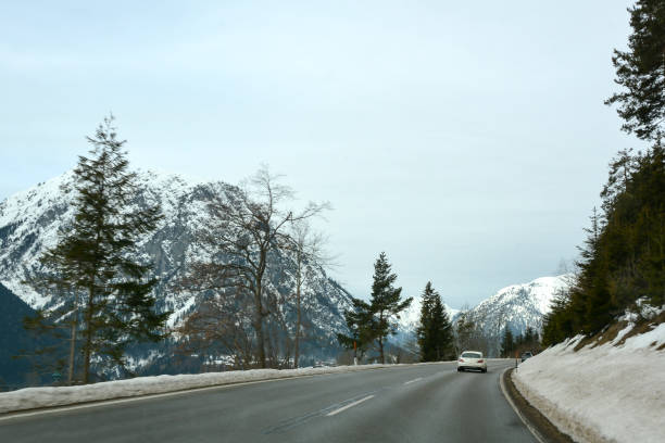 A winter road in perspective between small forested and snow-covered mountains under a clear blue sky. Cars drive in the distance