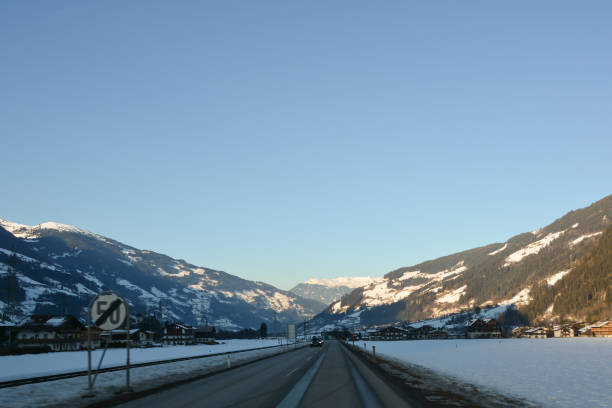 A winter road in perspective between small forested and snow-covered mountains under a clear blue sky. Cars drive in the distance
