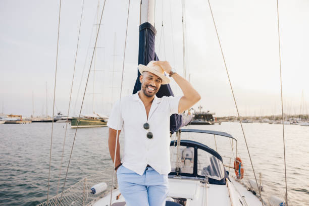 A cheerful man wearing a hat and casual attire stands on a sailboat. He's enjoying a sunny day at the marina, surrounded by calm waters and docked yachts.