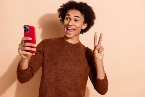 istock Young man with bright smile holding a red smartphone in hand, making a peace sign gesture against a beige background 2179717850