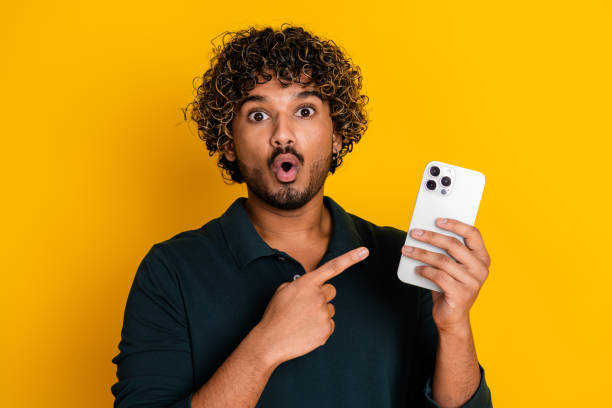 istock Young man with surprised expression pointing at smartphone against bright yellow background 2179720680