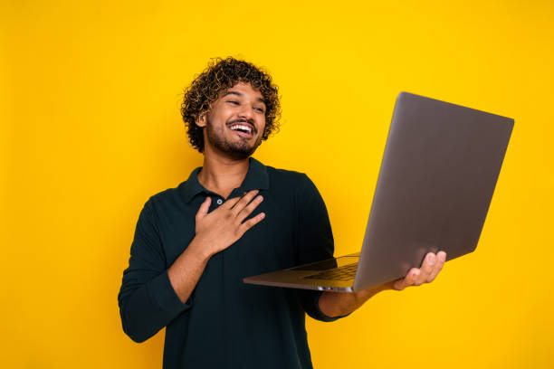 istock Young man smiling while holding  laptop against vibrant yellow background perfect for advertising and promotional concepts 2179720726
