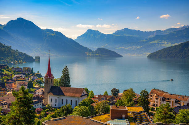 Weggis Village Overlooking Lake Lucerne and Swiss Alps A scenic view of Weggis, Switzerland, featuring a church with a red steeple, Lake Lucerne, and the surrounding Swiss Alps under a clear blue sky. lucerne switzerland stock pictures, royalty-free photos & images