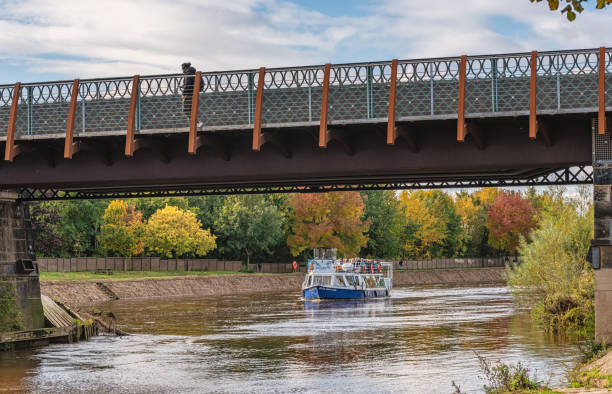 York, UK. October 10, 2024.  A tour boat packed with tourists approach a bridge over a wide river.  A pedestrian crossed the bridge and a sky with cloud is above.