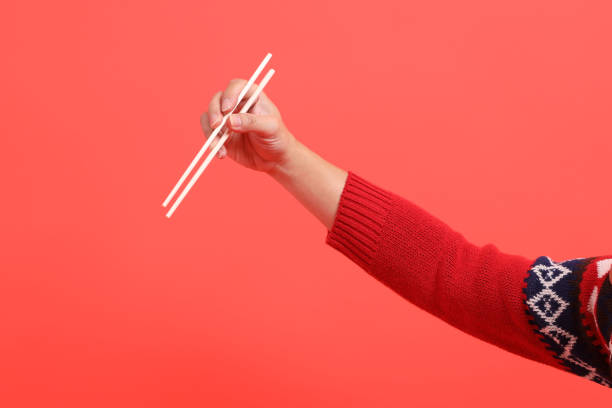 A close-up of a hand wearing a red cardigan holding up wooden chopsticks , isolated against a red background. Merry Christmas.