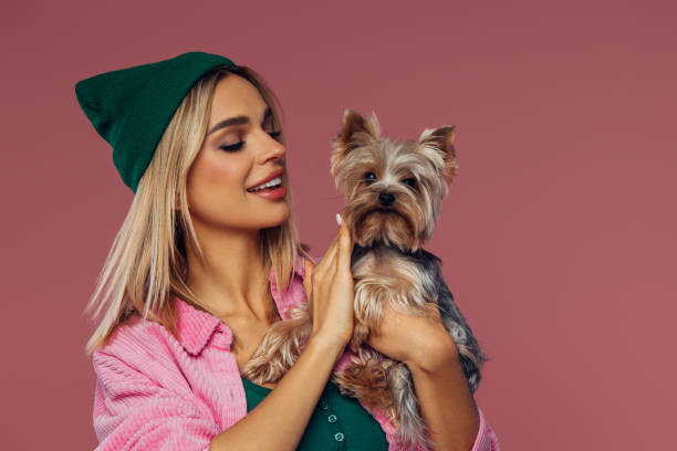 A heartwarming portrait of a young woman embracing her adorable Yorkshire Terrier. She exudes casual chic style in a green beanie and a pink corduroy shirt over a green top, perfectly complemented by her blonde hair. The soft pink background enhances the cozy and affectionate mood of the image. The woman gazes lovingly at her pet, who looks back with an equally endearing expression. This image is ideal for themes of friendship, pets, casual fashion, and warm, affectionate moments.