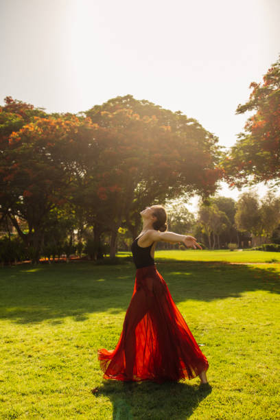 A woman gracefully dances in a sunlit garden, her red skirt billowing behind her as she moves. The vibrant greenery and sunlight cast a serene atmosphere, highlighting her lightness and joy. Dressed in a flowing red skirt and a black top, she embodies freedom, expression, and elegance, creating a captivating moment of movement and beauty. The warm sunlight filtering through the trees enhances the natural, outdoor setting.
