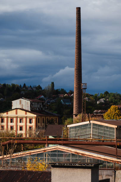 A view of aging industrial buildings with rusted metal pipes, glass windows, and a tall chimney, set against a backdrop of a rural landscape with houses and trees. The image captures the contrast between industrial decay and the surrounding nature, under a moody, overcast sky.