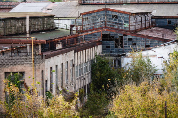 A view of aging industrial buildings with rusted metal pipes, glass windows, and a tall chimney, set against a backdrop of a rural landscape with houses and trees. The image captures the contrast between industrial decay and the surrounding nature, under a moody, overcast sky.