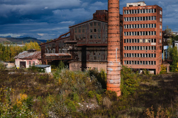 A vast, abandoned industrial complex featuring a tall brick chimney, rusted metal structures, and decaying buildings. The scene is set in an overgrown area with autumn foliage, under a moody sky, capturing the stark contrast between industrial decline and nature's encroachment.