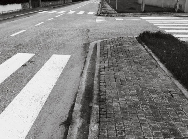 A black and white image of an urban road featuring several zebra crossings in close proximity. The paved sidewalk runs parallel to the road, with patches of grass visible near the curb. The street appears empty, with no vehicles or pedestrians present. The repetitive pattern of the zebra crossings adds a sense of geometric order to the scene, while the rough texture of the cobblestone sidewalk contrasts with the smooth road surface