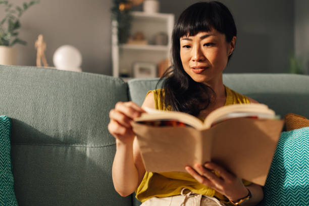 Focused asian woman flipping through book pages on comfortable sofa.