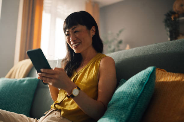 istock Asian woman smiling at smartphone while sitting on a comfy sofa. 2179848982