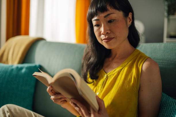 istock Asian woman deeply engrossed in reading a book on comfortable couch. 2179848995