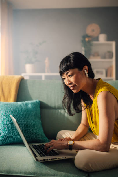 istock Asian woman on a sofa working on a laptop, surrounded by cushions. 2179849030