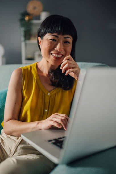 istock Asian woman working on laptop, thoughtful expression, teal couch modern 2179849038