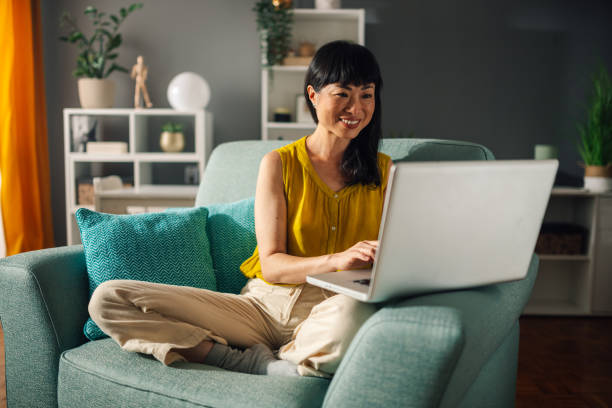 istock Asian woman cross-legged on couch using laptop in cozy home setting 2179849061