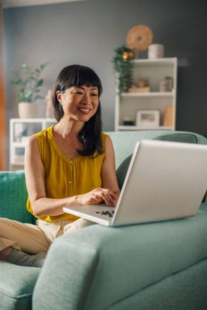 istock Focused asian woman typing on a laptop while sitting on a comfy couch. 2179849067