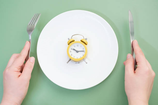 istock Female hand holding a fork and knife next to an empty plate with alarm clock on a green background close-up top view. 2179855241