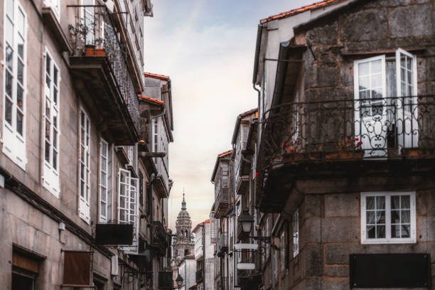 Narrow European street with traditional stone buildings and wrought iron balconies, leading towards a historic cathedral tower. Soft evening light adds warm tones to the scene