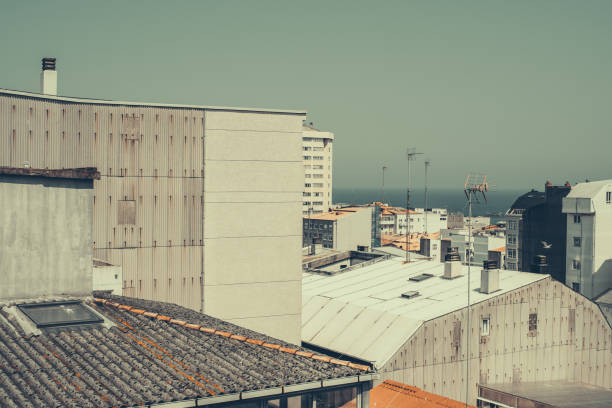 Urban rooftops and facades of industrial buildings in the foreground with sea view in the background. Antennas and ventilation chimneys are visible on top of the structures