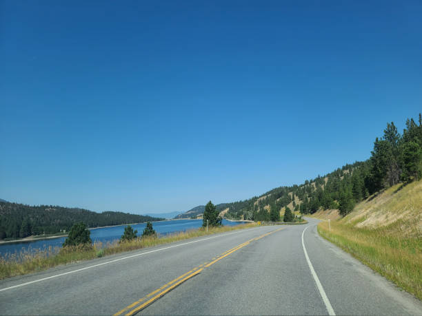 A scenic road curves alongside a tranquil lake with forested hills under a clear blue sky.