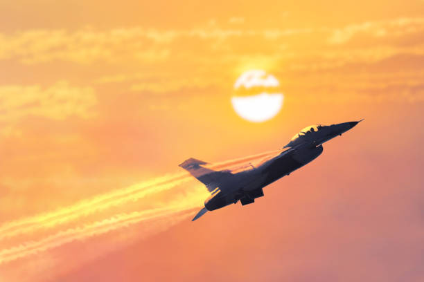 A military jet flies through burnt cloudy skies with the sun in the background