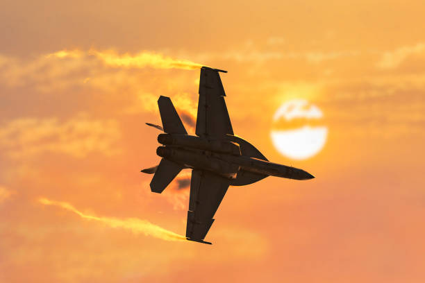 A military jet flies through burnt cloudy skies with the sun in the background