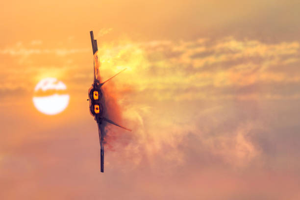 A military jet flies through burnt cloudy skies with the sun in the background