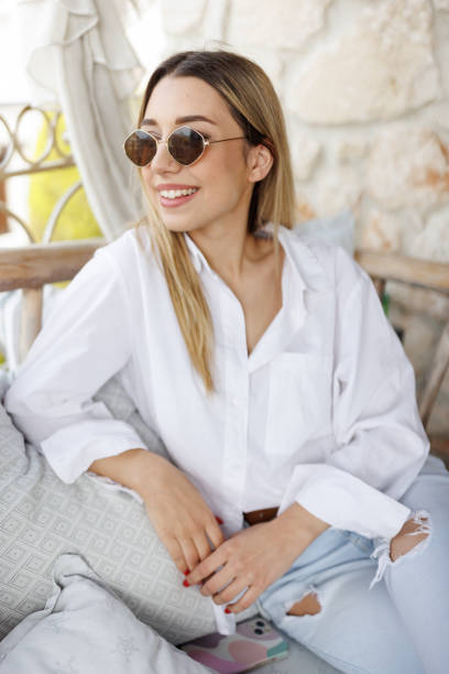 Smiling young woman with sunglasses sitting on balcony sofa and enjoying sun