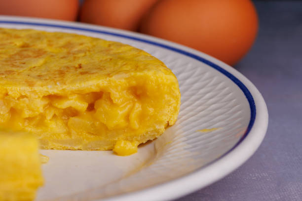 Spanish omelette with a missing slice, along with bread, eggs, and a red checkered napkin, atop a blueish concrete surface