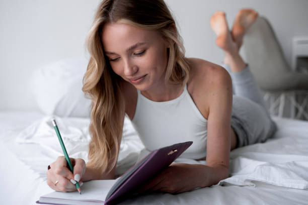 istock Pensive young woman lying on bed with serious expression writing on journal. 2179930391
