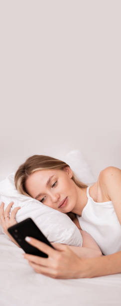 istock Cute woman awake lying in comfortable white bed checking mail on smartphone 2179930451
