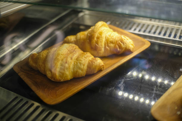 Two freshly baked croissants displayed on a wooden tray inside a glass bakery case. Their golden, flaky crusts are highlighted by the warm lighting, enticing customers.