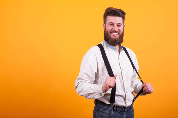 Hipster bearded man pulling his lederhosen over yellow background, Man smiling. Looking at the camera. Stylish man.